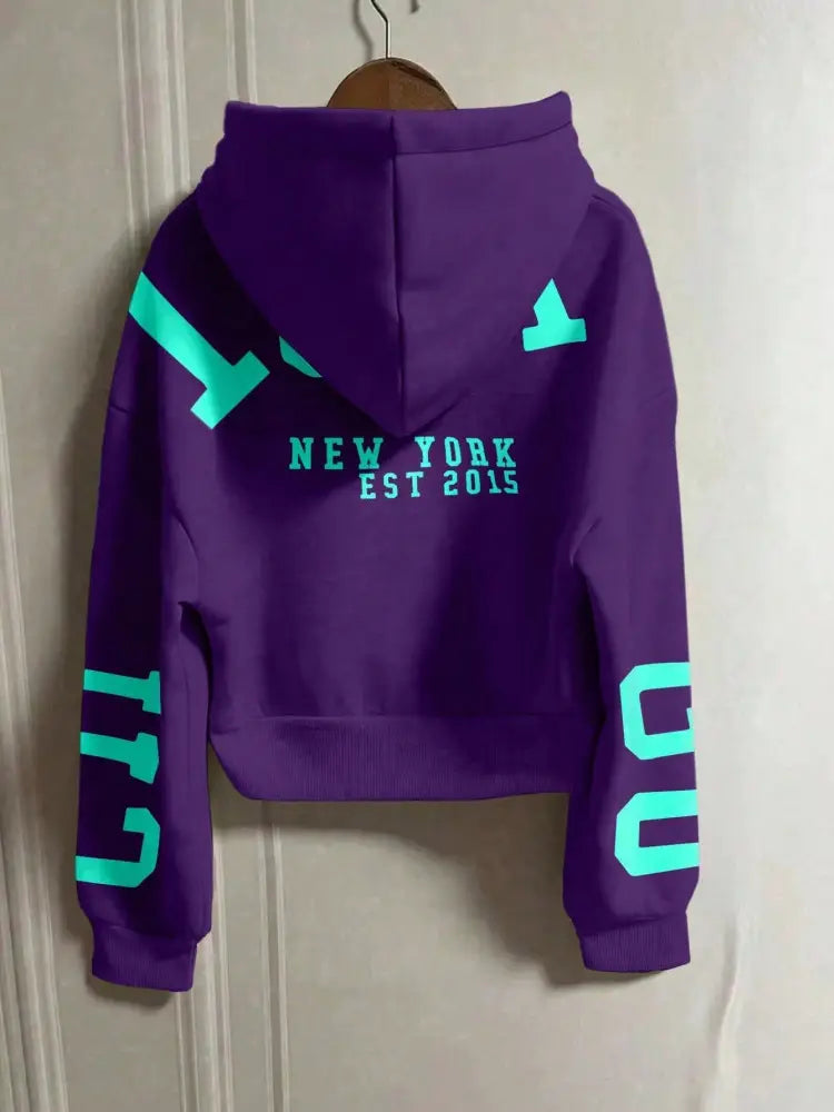 Face arrière d’un sweat-shirt à capuche zippé imprimé graphique violet court, avec des lettrages géométriques vert néon et « New York Est 2015 » imprimé sur l’ensemble du dos.