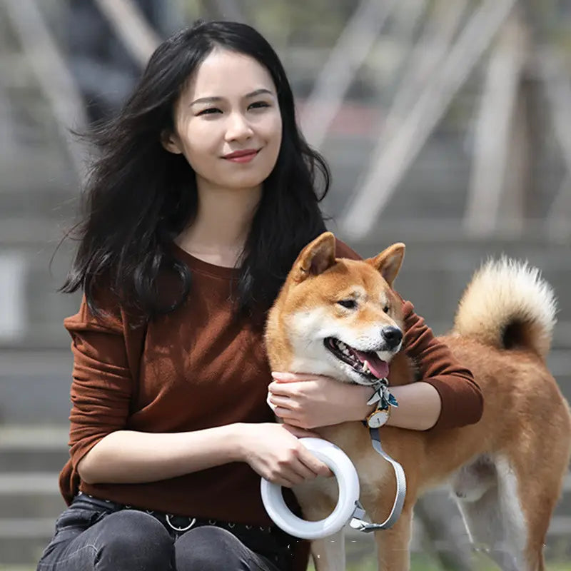 A smiling woman with long dark hair sits outdoors holding a cheerful reddish-brown shiba inu dog using a retractable hands free dog leash with led light.