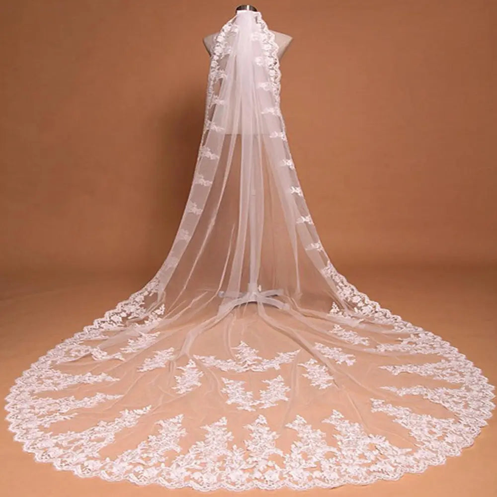A long, flowing white lace cathedral wedding veil with intricate floral embroidery along its edges and a wide decorative hemline, featuring a comb for secure attachment.