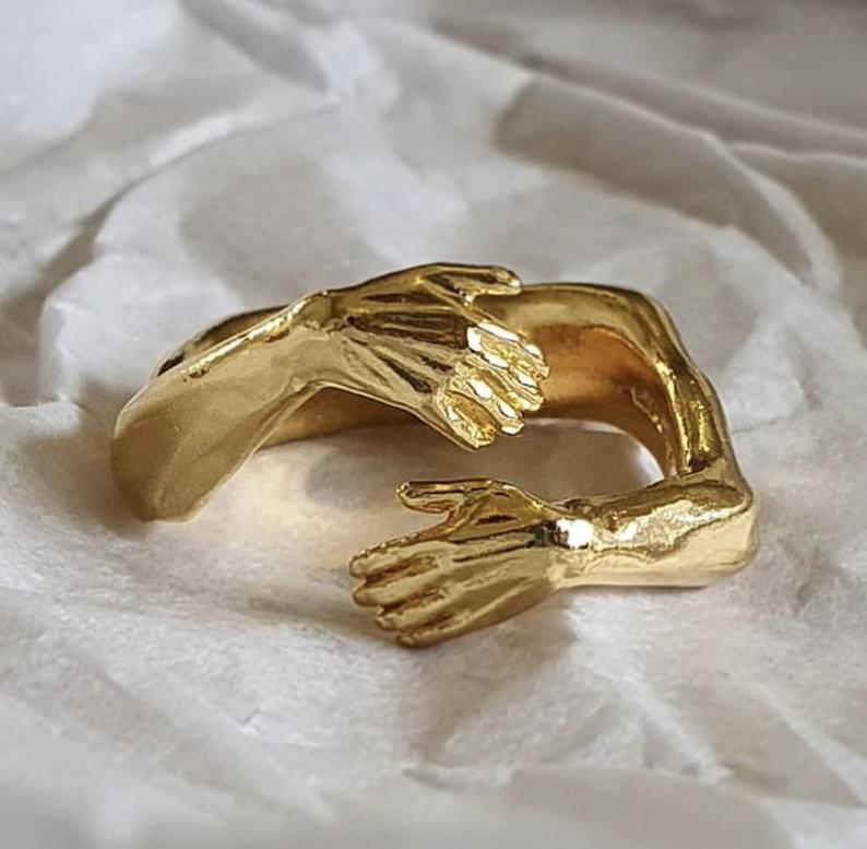 Adjustable silver ring with gold-toned sculpted hands embracing each other gently featuring a red gemstone.