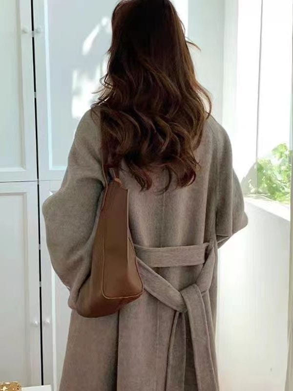 Eine Schultertasche aus beigem Leder mit einer strukturierten, minimalistischen Gestaltung und passendem Riemen, die zusammen mit einem Kaschmir-Wollmantel mit doppelter Wollseite und gegürteter Passform präsentiert wird.