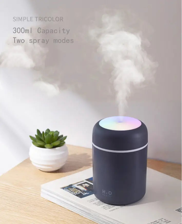 A sleek matte black ultrasonic mini humidifier with a glowing multicolored rainbow LED light top emitting mist and featuring the h2o logo on its side with a 300ml capacity.