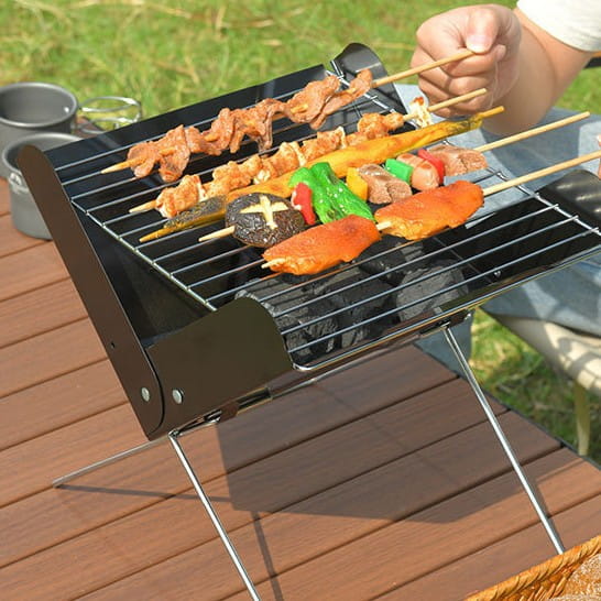 Black foldable portable charcoal grill with stainless steel legs and grilling rack cooking skewers of meat and vegetables for outdoor camping use.