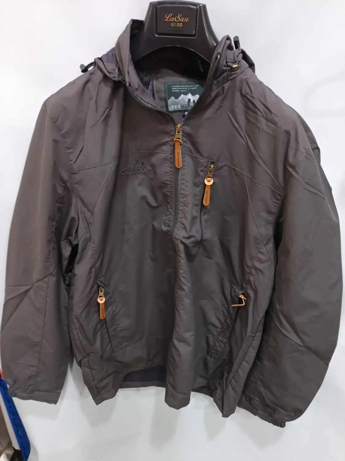 Chaqueta de senderismo con capucha impermeable color gris oscuro, con tiradores de cremallera color camel colgando de una percha negra con marca "lasian"