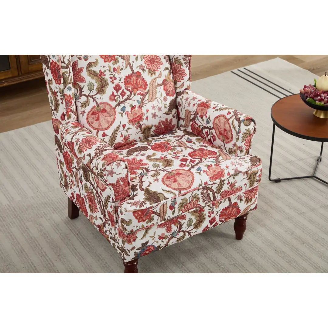 Una cómoda silla tipo ala con respaldo capitoné, tapizada en un tejido floral vibrante con flores rosadas rojas y marrones sobre un fondo blanco, con patas de madera retro.