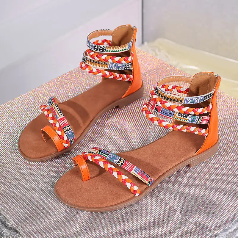 Orange flat sandals with colorful braided ankle straps featuring bohemian ethnic geometric patterns.