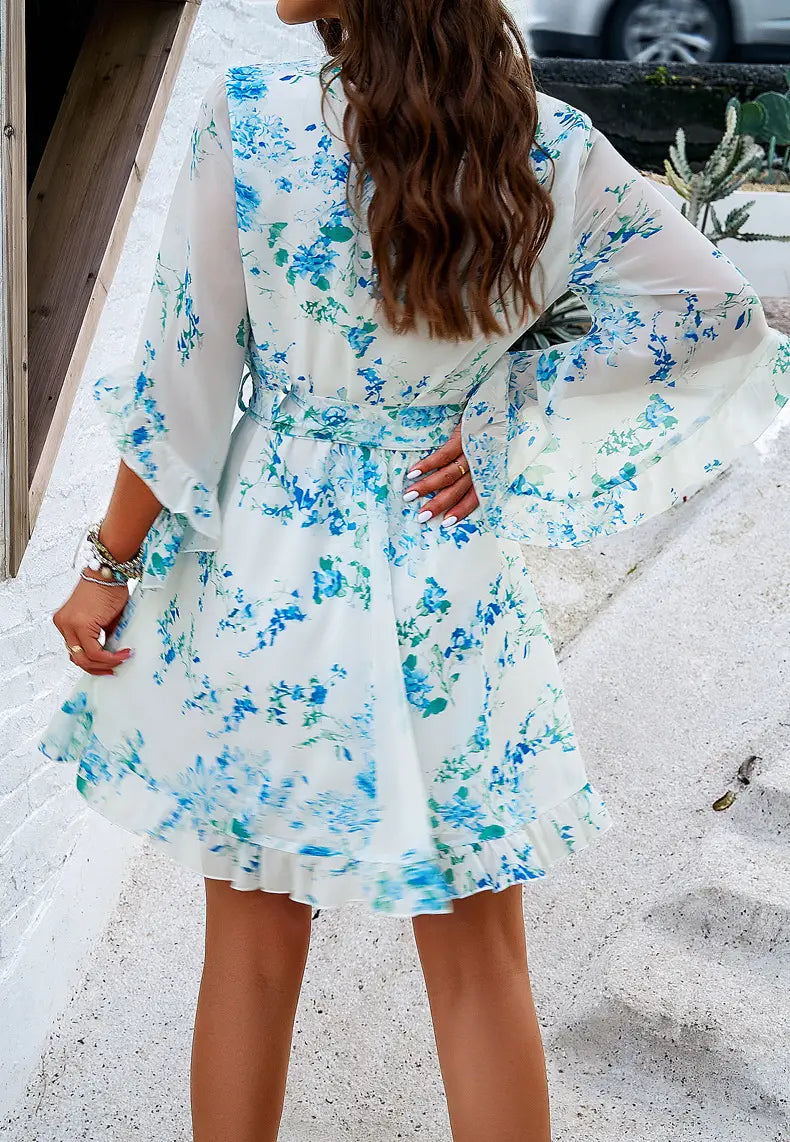 Women’s floral v-neck ruffle dress with waist tie in white fabric featuring blue and green blossoms, bell sleeves, and a ruffled hem.