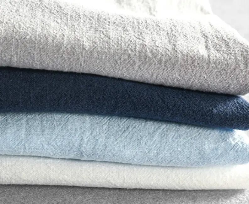 Pile de chemises en lin pour homme repliées dans des tons gris doux, bleu marine clair et blanc, mettant en avant des hauts d’été décontractés et respirants.