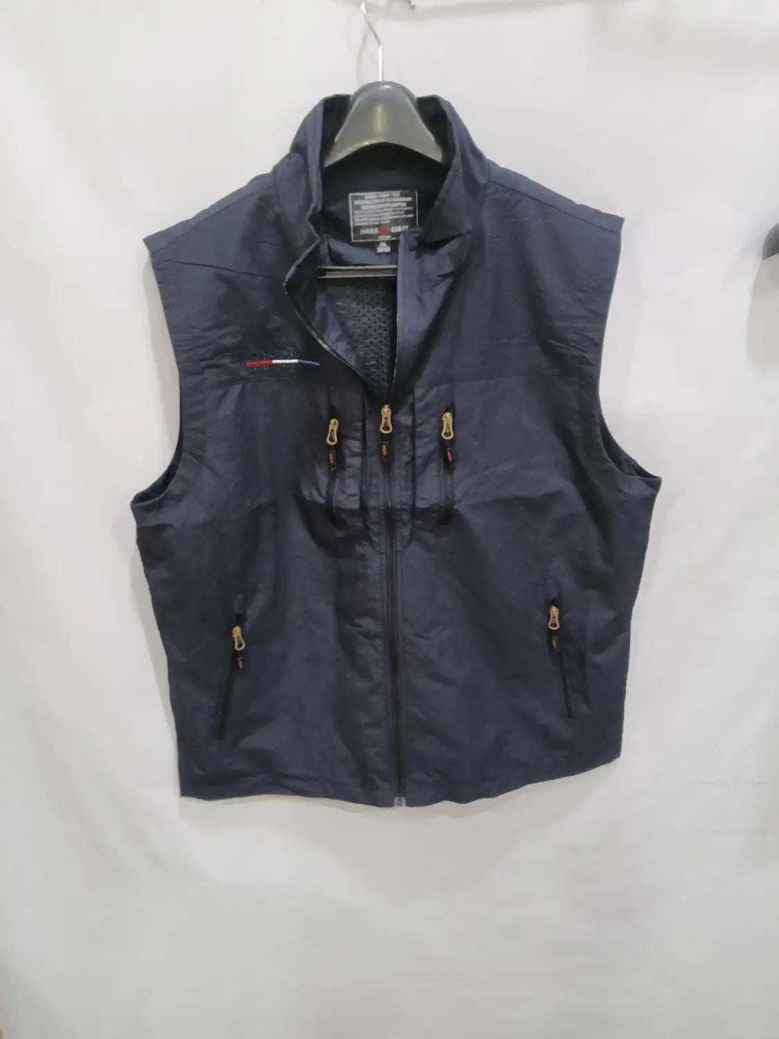 Chaleco utilitario con capucha sin mangas para hombre en azul marino oscuro, con cremalleras de tono dorado y un logotipo sutil rojo, blanco y azul en el lado izquierdo del pecho.
