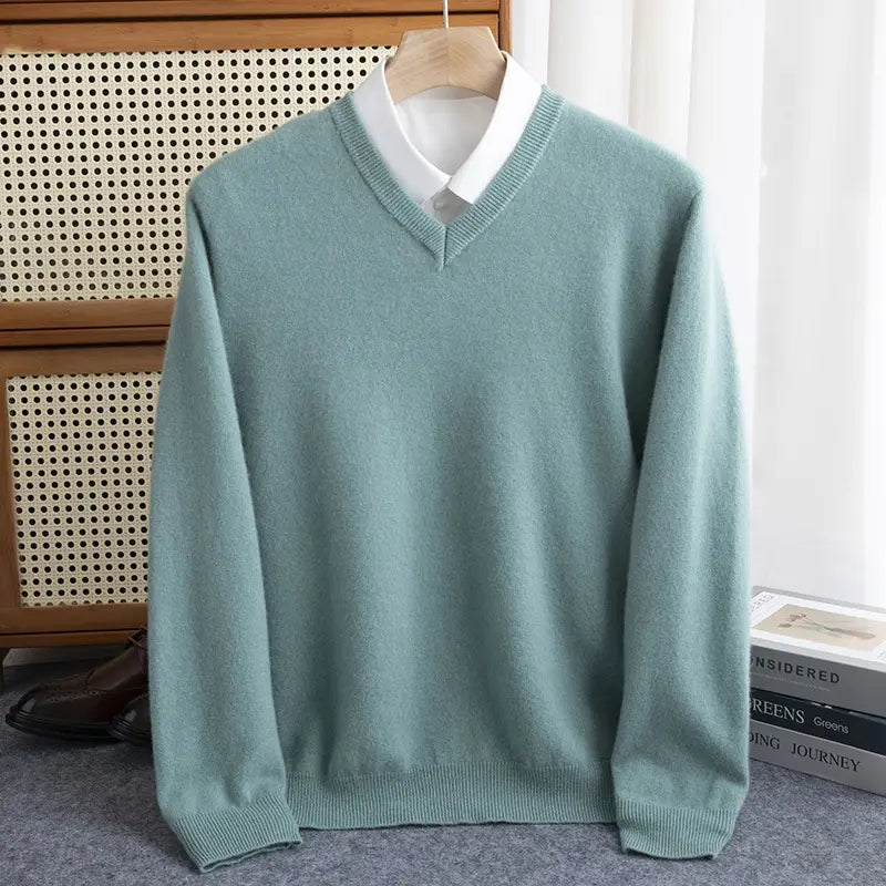 Ein weicher Wollstrickpullover in Salbeigrün für Herren mit V-Ausschnitt, gerippter Struktur und fertig ausgearbeiteten Bündchen, auf einem Holzkleiderbügel für den Winter gezeigt.