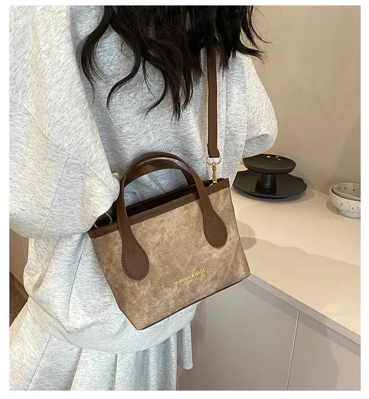 A taupe canvas retro bucket bag mini tote with brown leather handles and trim featuring a subtle embossed brand logo on the front.