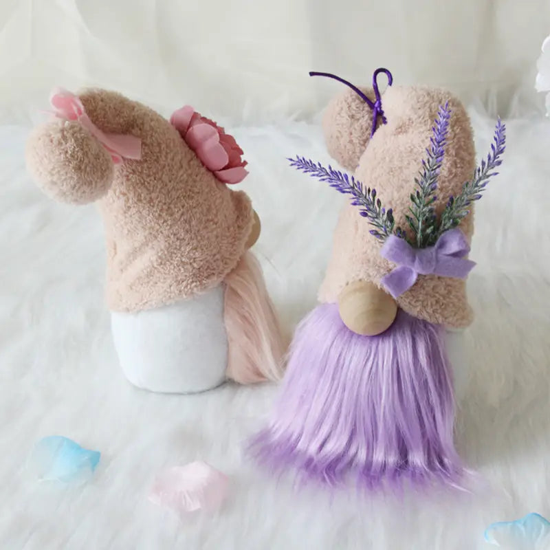 Two plush gnome dolls in pink and lavender holding a pink flower and lavender sprigs with a purple bow displayed as desktop ornaments.