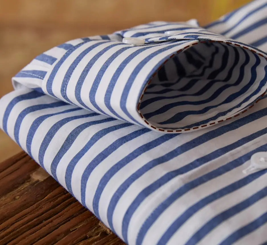 Ein ordentlich gefaltetes Slim-Fit gestreiftes Baumwoll-Hemd langarm für den Pendlerbereich für Herren mit kräftigen blau-weißen Vertikalstreifen und einer geknöpften Manschette mit kontrastierenden braunen Ziernähten.