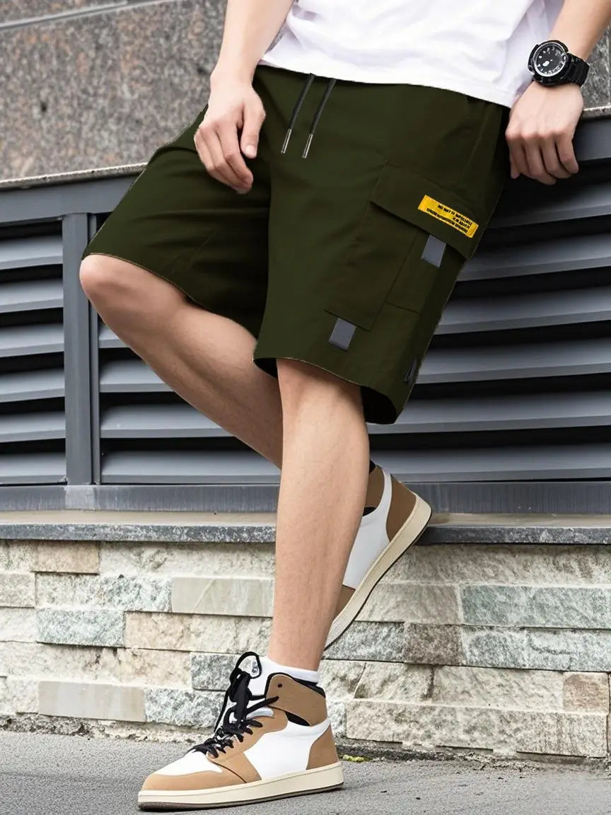 Shorts cargo utilitaires pour homme vert olive, avec une étiquette jaune et des empiècements réfléchissants gris, munis de poches à cordon de serrage adaptés à une tenue d’été.
