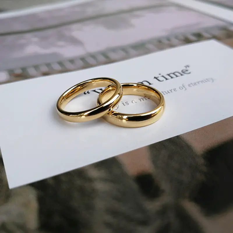 Two polished gold tungsten steel wedding bands with a smooth rounded design rest on a white card for couples.