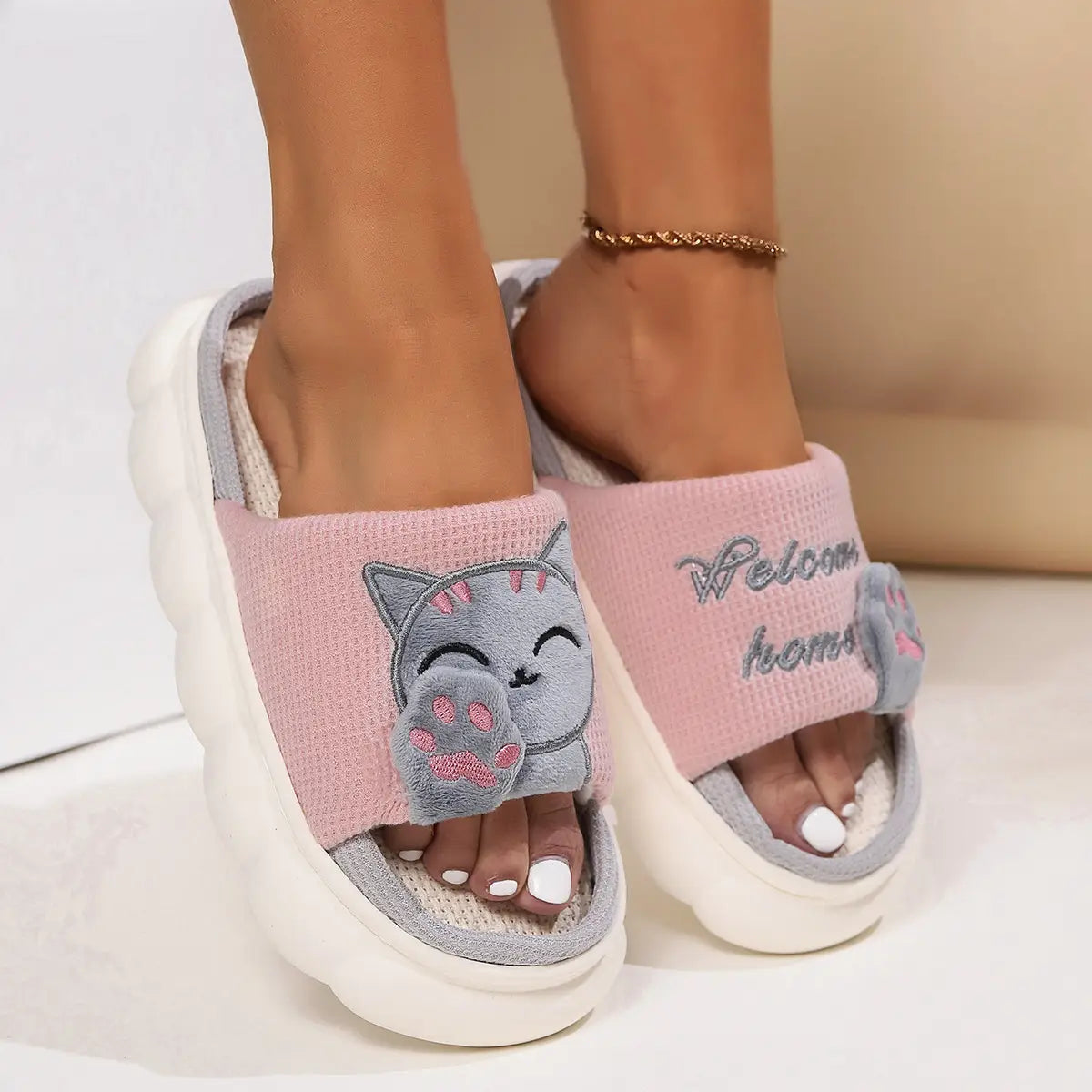 Pink and gray plush embroidered cat cartoon unisex anti slip slide slippers with a cute cat face, pink paw details, and the phrase welcome home stitched on the side.
