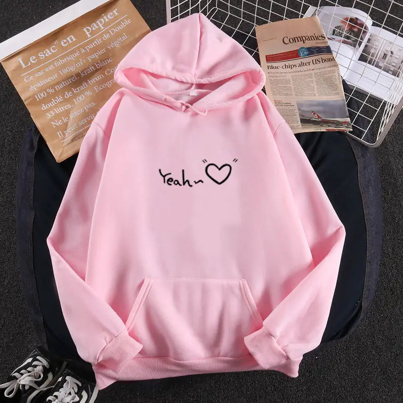 Sudadera con capucha aislada y forro polar rosa suave, con un gráfico de corazón 'yeah' negro en el pecho, que incluye capucha con cordón y bolsillo delantero.