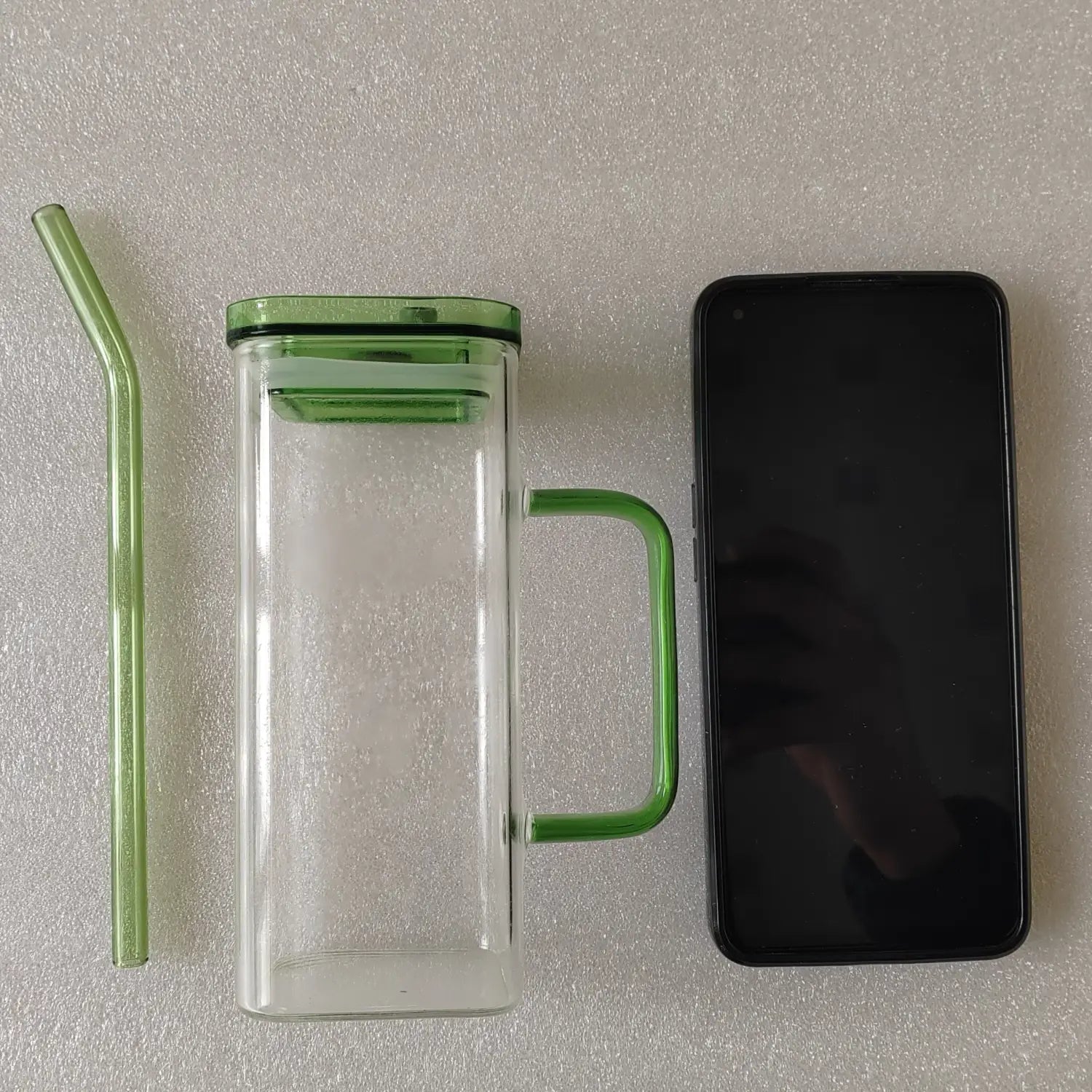 Gobelet en verre transparent à double paroi avec poignée de couvercle en plastique vert et paille incurvée assortie, présenté de profil dans une tasse carrée en verre de 12 oz avec couvercle et paille.