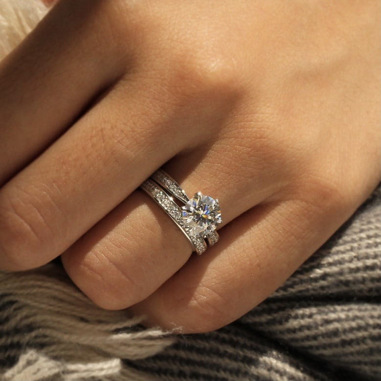 A sparkling round cut zircon wedding ring set in white gold finish with a central round-cut stone and pavé-set side stones worn on a finger.