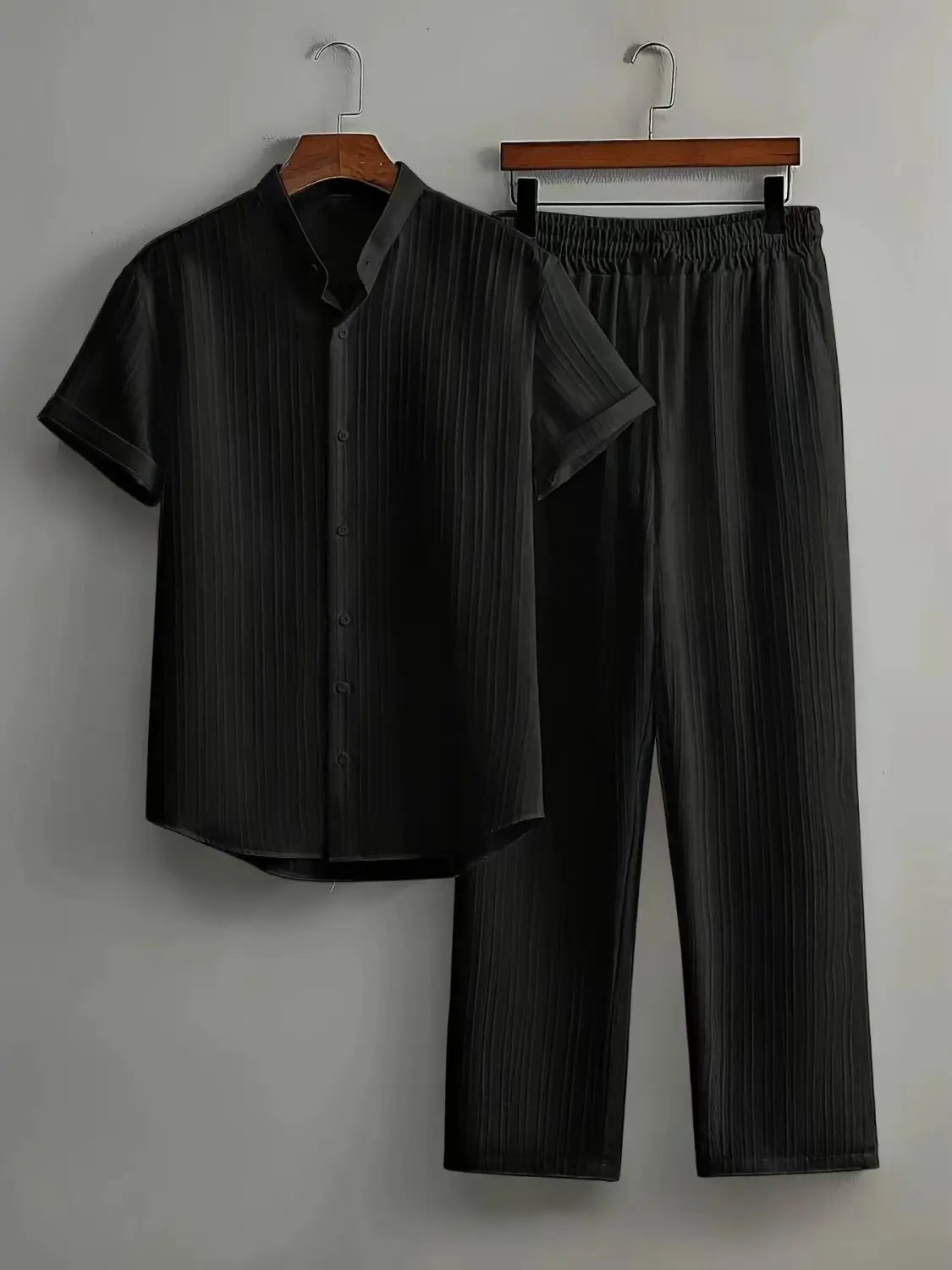 Black mens pleated short sleeve button-up shirt with vertical pleats and matching black trousers hanging on wooden hangers.