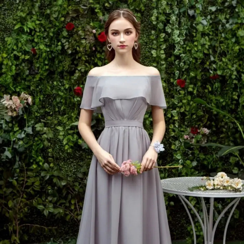 Off-the-shoulder gray chiffon floor length bridesmaid gown with a flowing design, fitted waist, and ruffled sleeves.