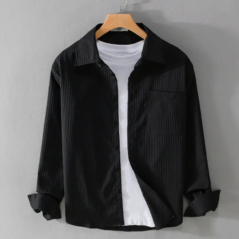 Chemise homme décontractée à manches longues en coton côtelé noir, avec un col classique et une poche poitrine, présentée sur un cintre en bois au-dessus d’un t-shirt blanc.
