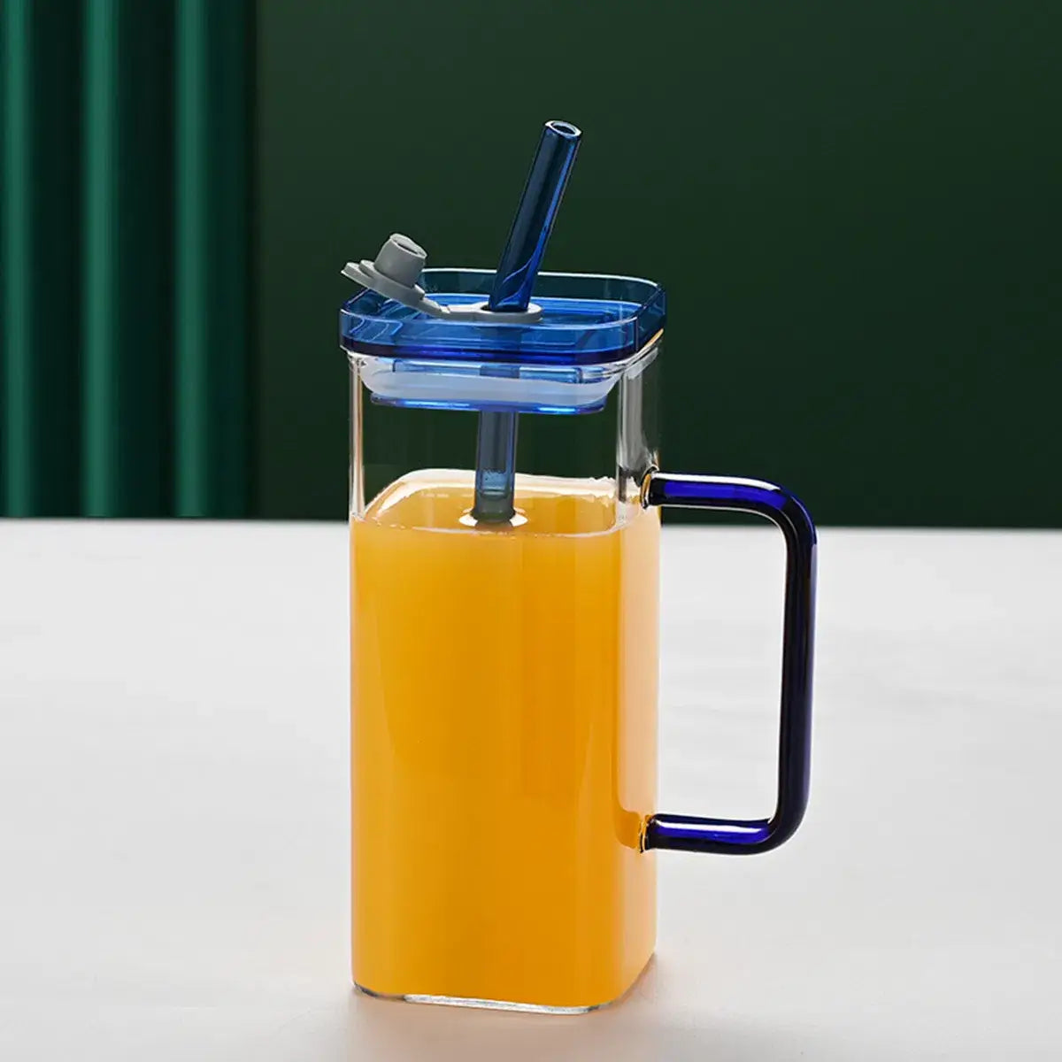 Carafe en verre transparent avec poignée et couvercle bleus, remplie de jus d’orange, à côté d’une tasse carrée en verre avec couvercle et paille de 12 oz.