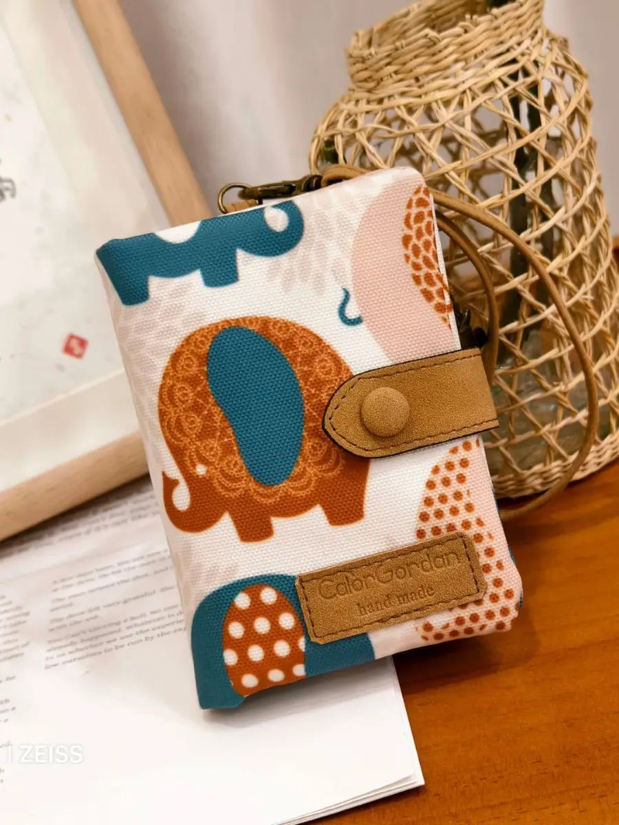 A whimsical patterned canvas tri fold multi card wallet featuring a playful teal and orange elephant design on a white background with tan leather straps and a branded tag.