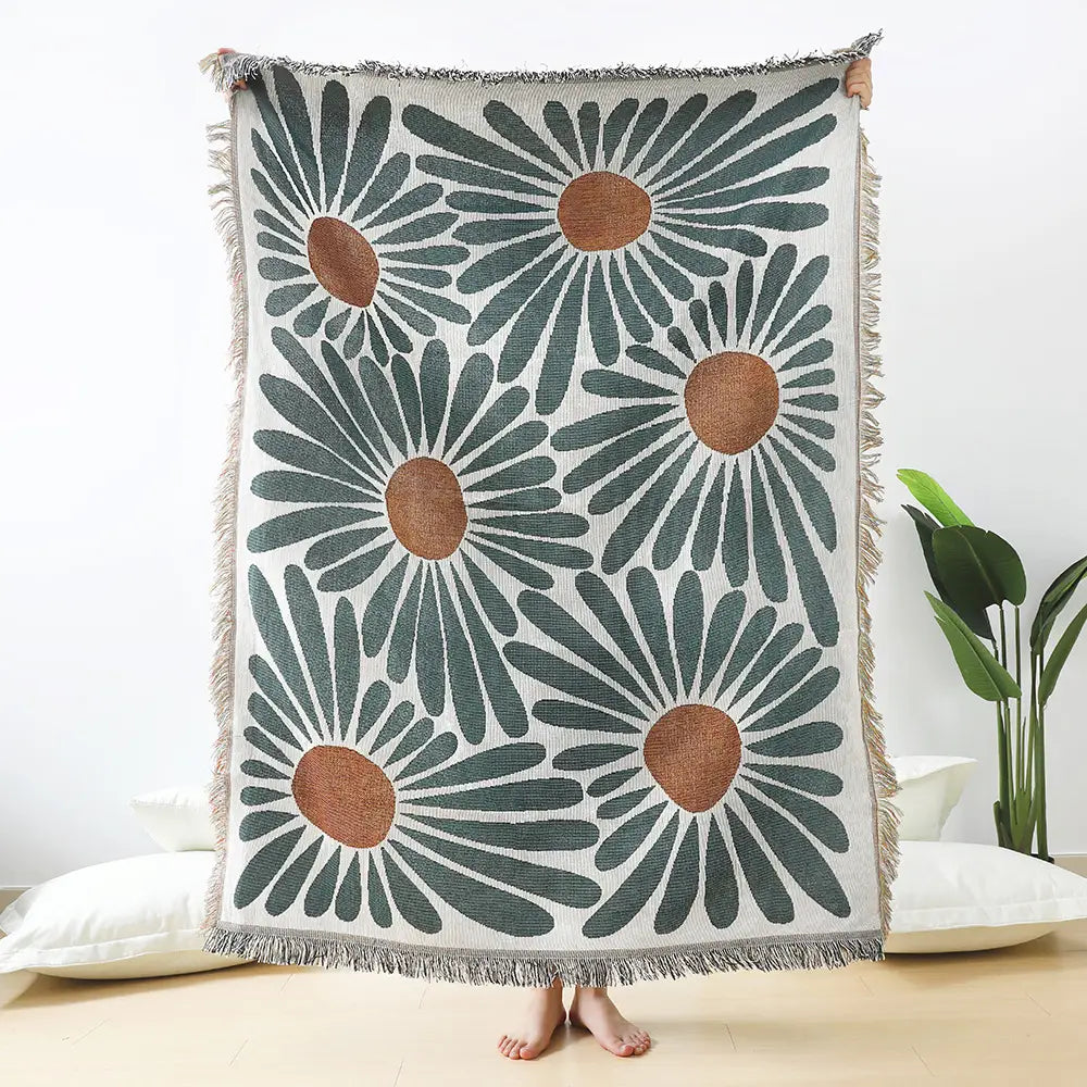 A cozy woven cotton throw blanket in cream featuring a bold pattern of oversized daisies with dark green petals and warm brown centers framed by textured fringe along all edges.