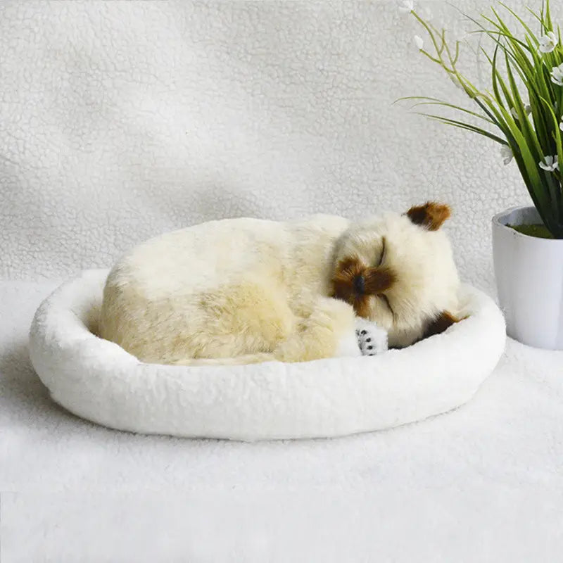A realistic breathing plush cat figurine in cream color with brown accents resting peacefully in a soft white circular pet bed.