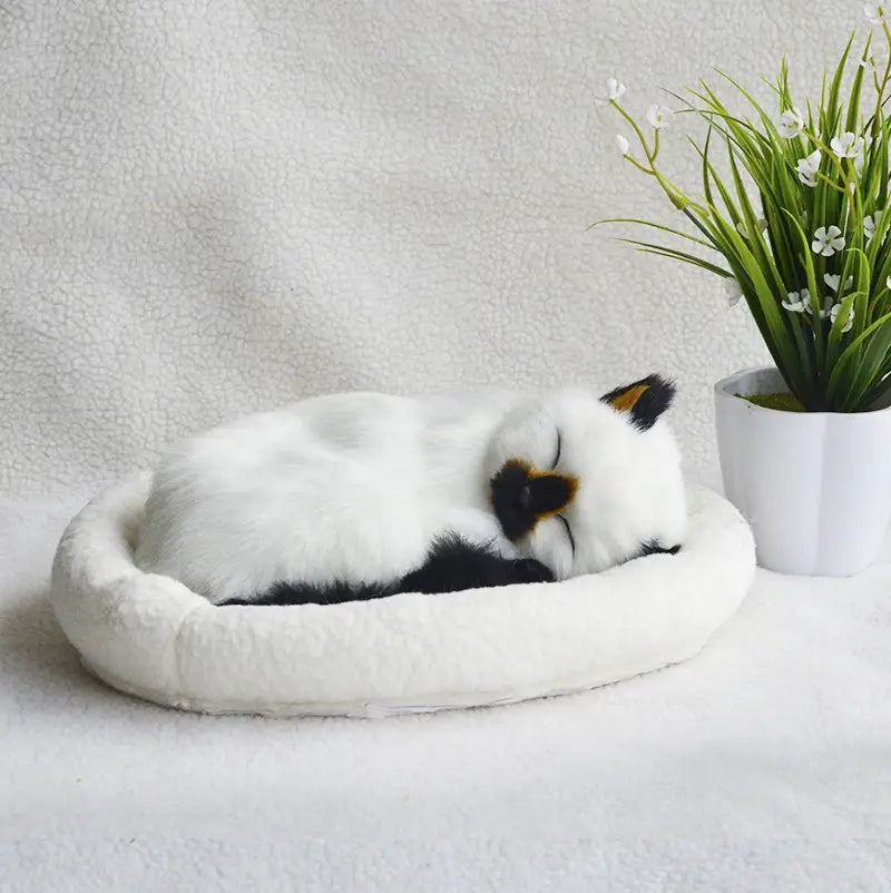 A plush white and black realistic breathing cat figurine with a soft fuzzy texture curled up asleep in a matching circular pet bed.