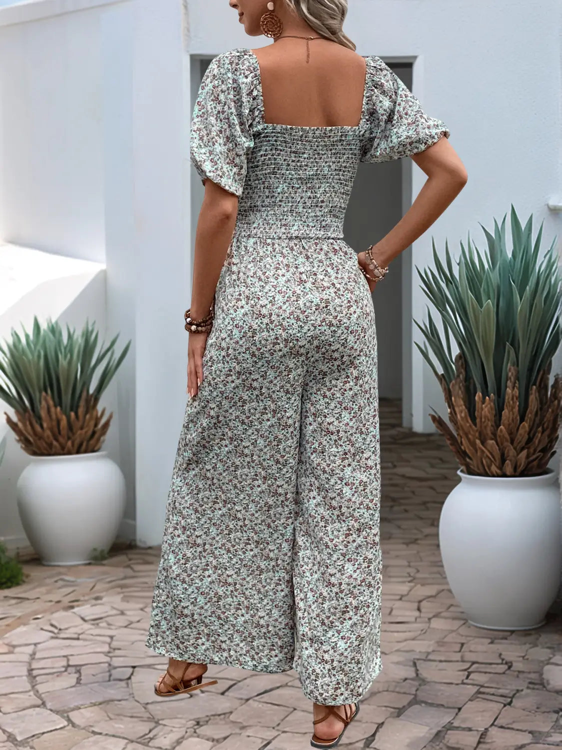 女性が、柔らかなグリーンとホワイトのトーンで流れるようなフローラルのパフスリーブとワイドレッグのジャンプスーツを、軽量な生地で身に着けています。