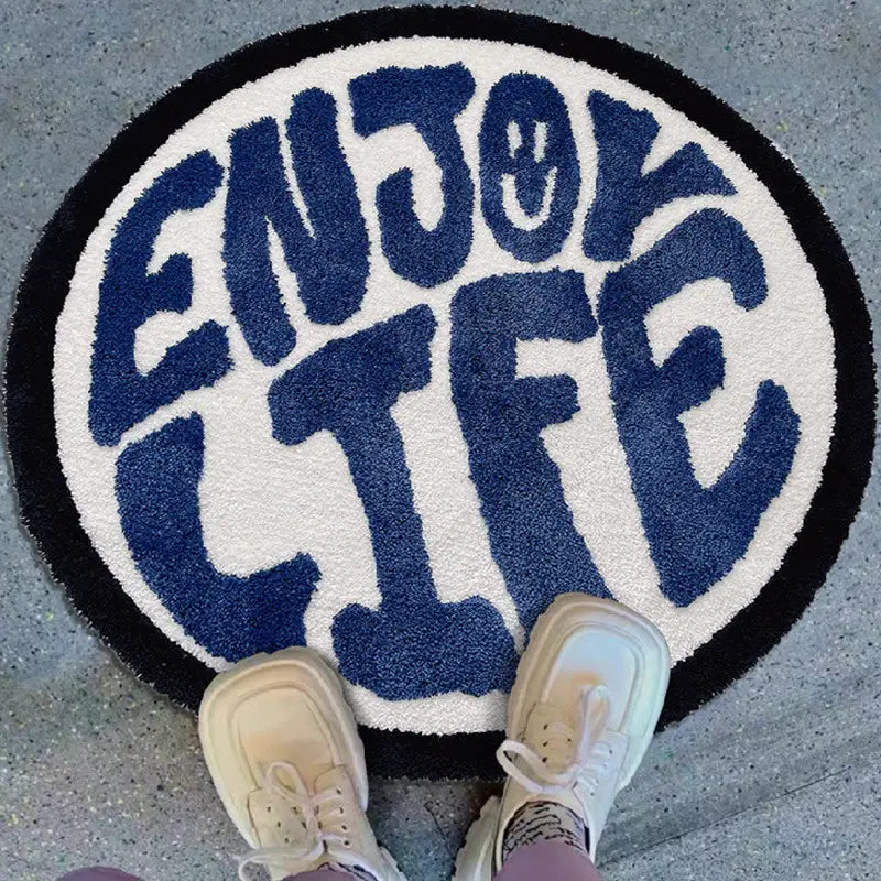 Round plush rug with enjoy life in bold navy blue lettering on a white background framed by a black border.