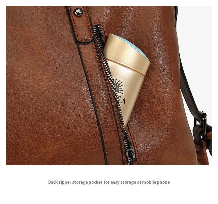 Gold-colored cylindrical travel mug with sunburst design and sa engraving in the back zipper pocket of the back side of a large capacity leather tote bag with shoulder strap.