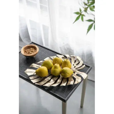 a black and white striped leaf-shaped serving tray holding yellow pears as part of gold monstera leaf wall decor and serving tray collection