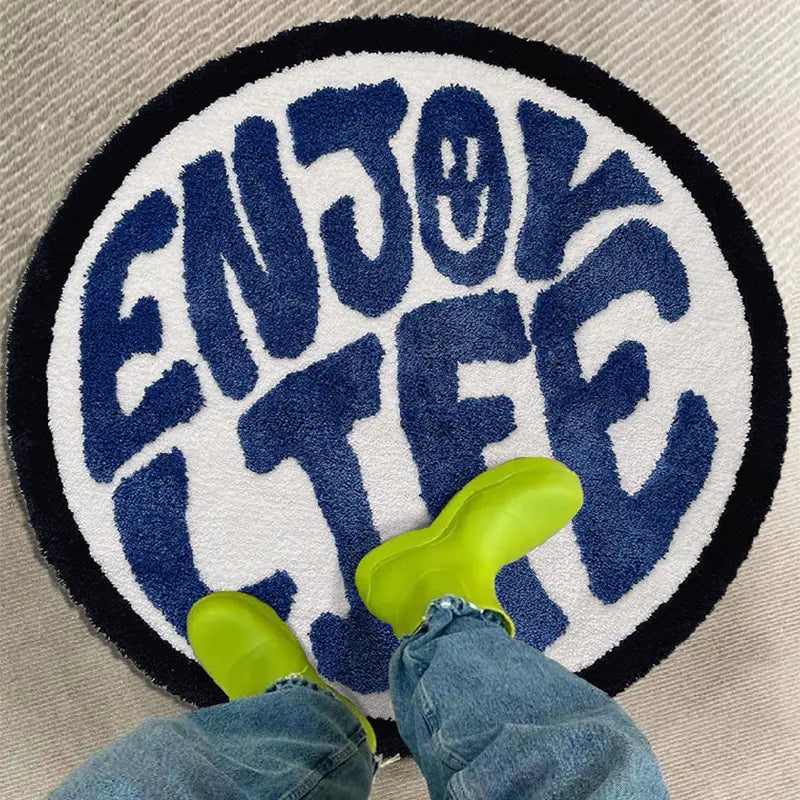 Circular plush rug featuring enjoy life minimalist print in bold navy blue lettering on a white background with a black border.