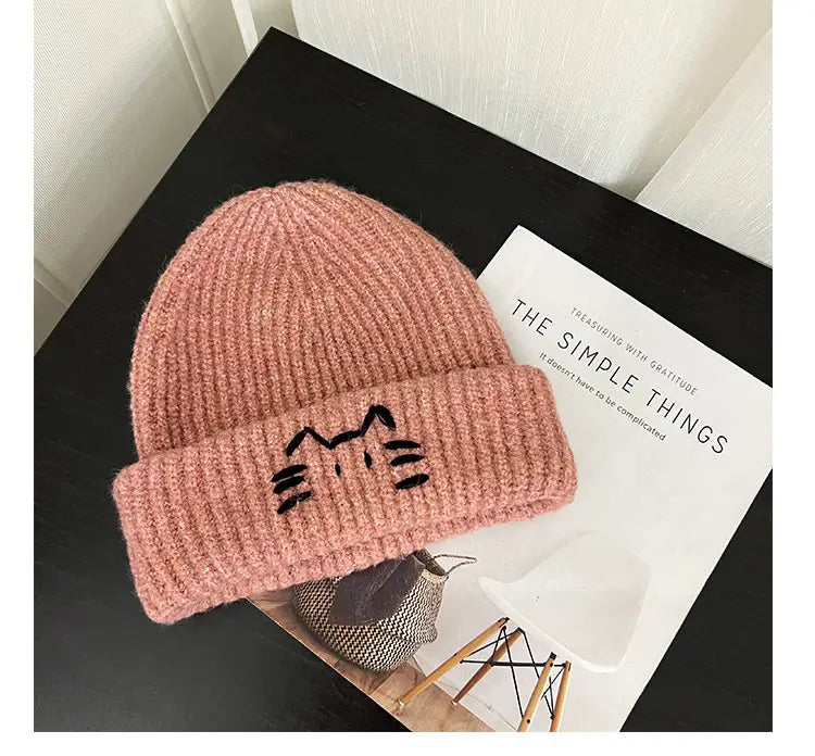 A soft pink knitted beanie hat with a black embroidered cat face on the folded brim.