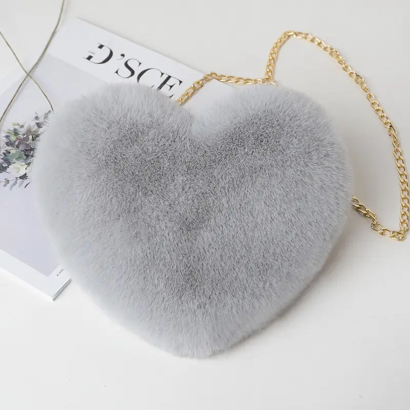 A soft light gray heart-shaped crossbody bag made of plush faux fur with a delicate gold chain strap.