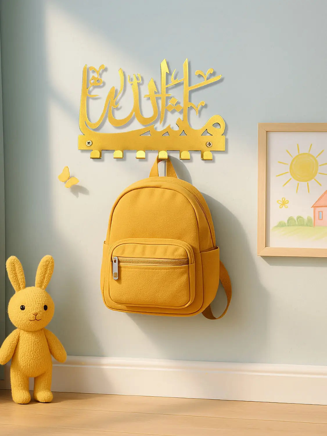 a mustard-yellow canvas backpack with a front zipper pocket and adjustable straps hangs from a golden metallic mashallah arabic calligraphy metal wall hook rack mounted on the wall