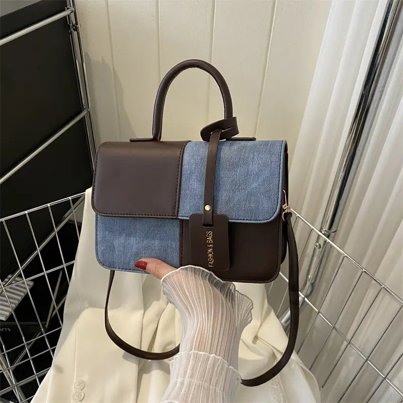 A stylish two tone denim patchwork crossbody bag featuring denim blue and dark brown leather with a top handle, adjustable shoulder strap, and a small gold toned tag.