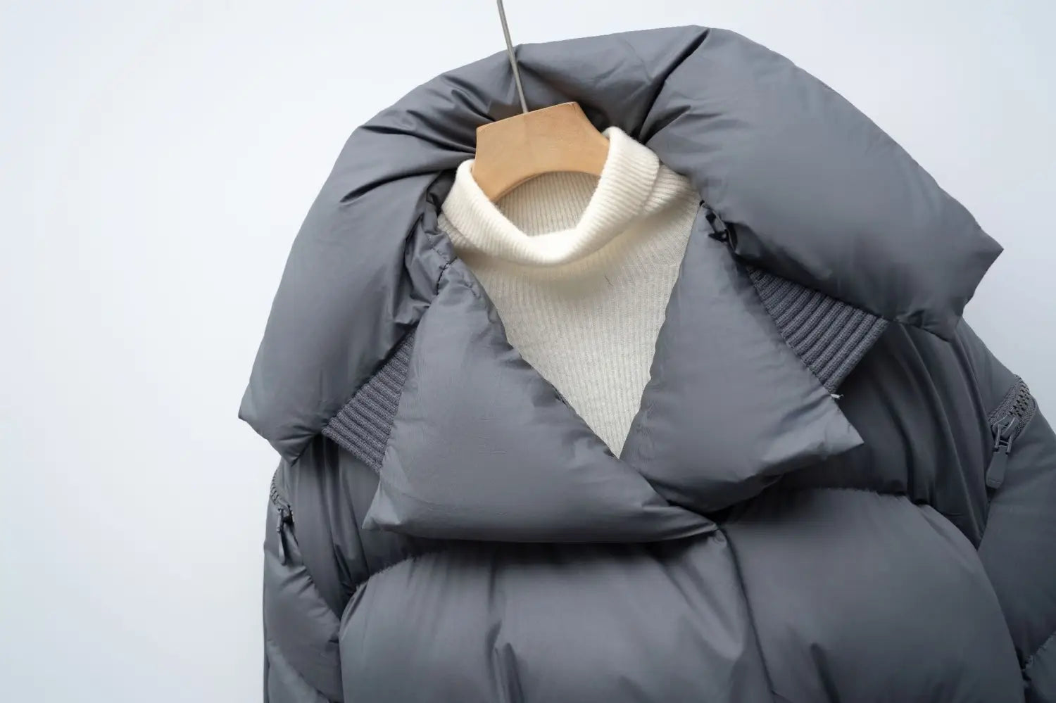 Gray down jacket patchwork with quilted design and ribbed trim hanging on wooden hanger over cream-colored turtleneck in Korean style large lapel.