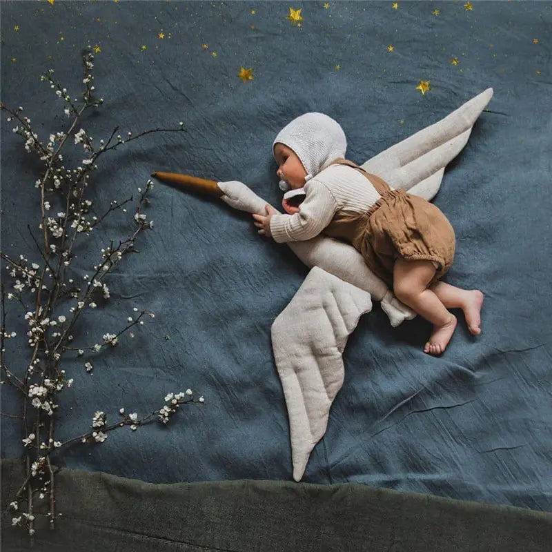 Un bébé habillé d’un bonnet en maille blanche et de salopettes beiges, couché sur un oreiller d’animal en peluche souple en forme de cygne blanc, avec des ailes souples.