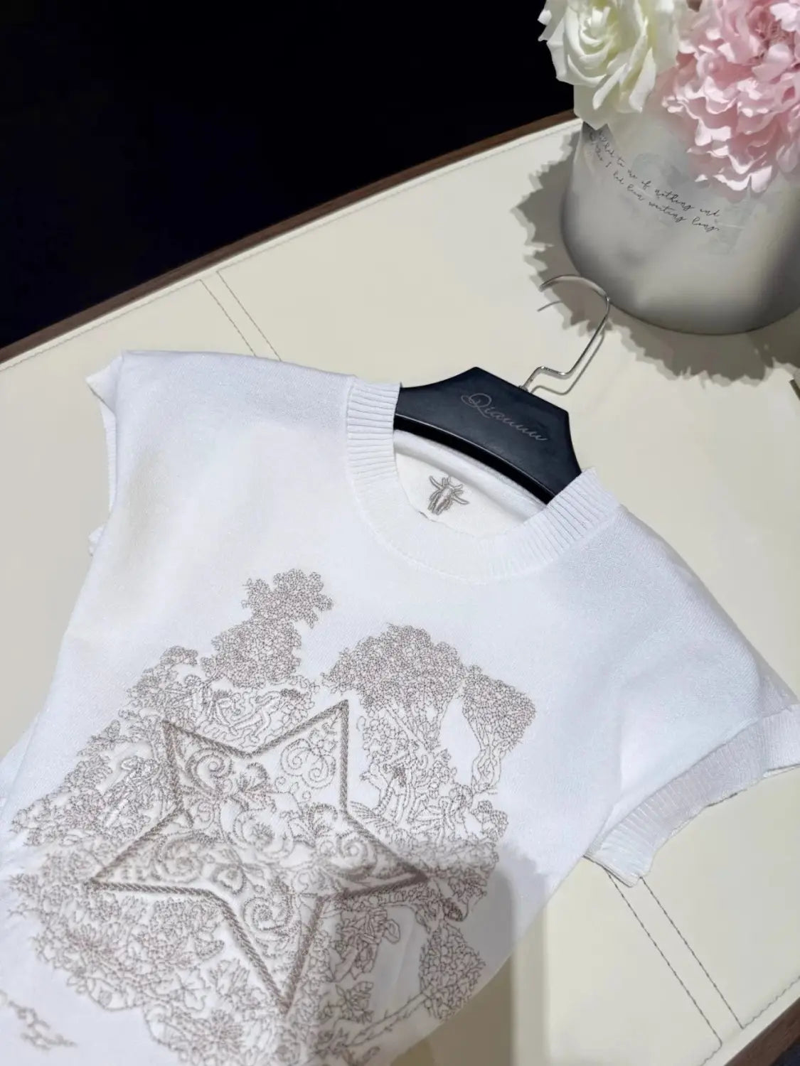 Weißes ärmelloses Strick-T-Shirt mit aufwendiger silberfarbener Stickerei, die Sterne- und florale Muster bildet, auf einem schwarzen Bügel präsentiert; mit elegantem Rundhalsausschnitt und beigen kurzen Ärmeln.