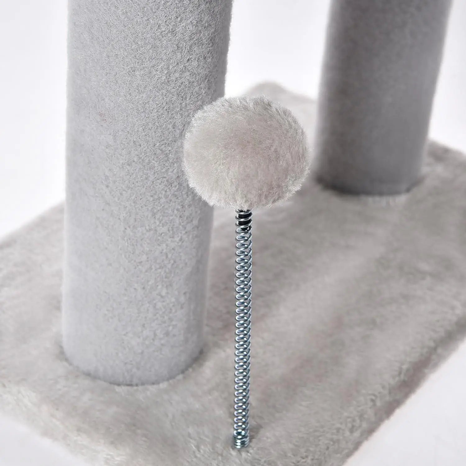 a fluffy white pom-pom attached to a metallic spring resting on the gray carpeted base of a gray cat tree with tunnel and scratching post