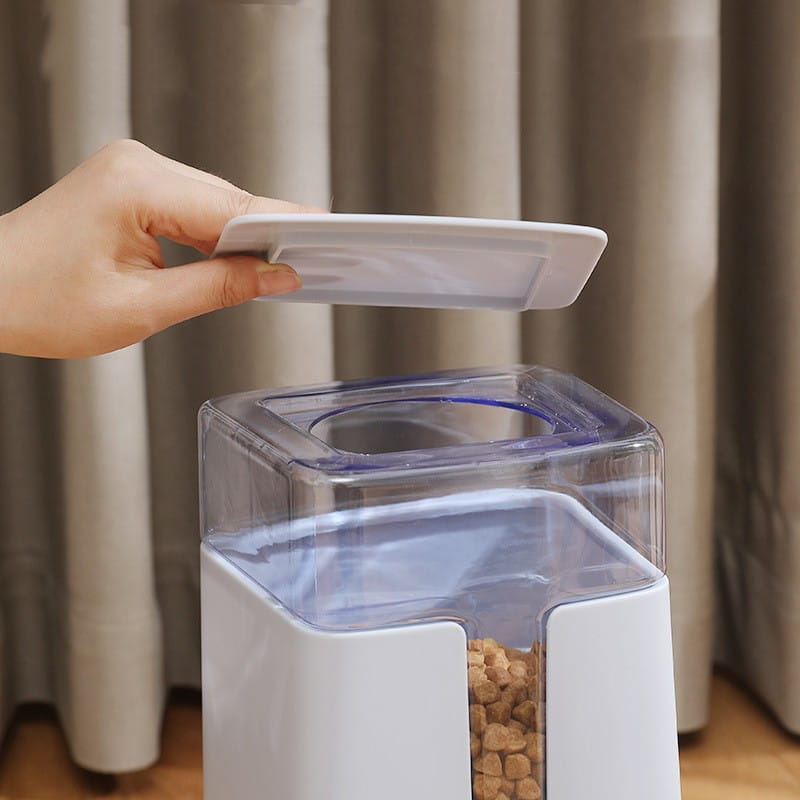 Automatic pet feeder and water dispenser in white plastic with a transparent top and blue rim containing brown kibble.