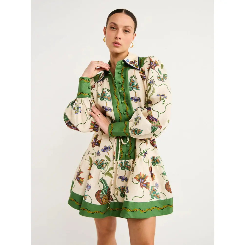 A woman wearing a cream-colored botanical print puff sleeve midi dress with an intricate green and purple botanical pattern and bold green contrast trim on the collar, cuffs, hem, and waistband.