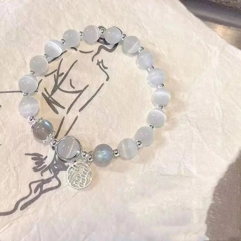 A delicate crystal butterfly cat’s eye bead bracelet featuring translucent light gray beads and a silver-toned circular charm with an engraved design.