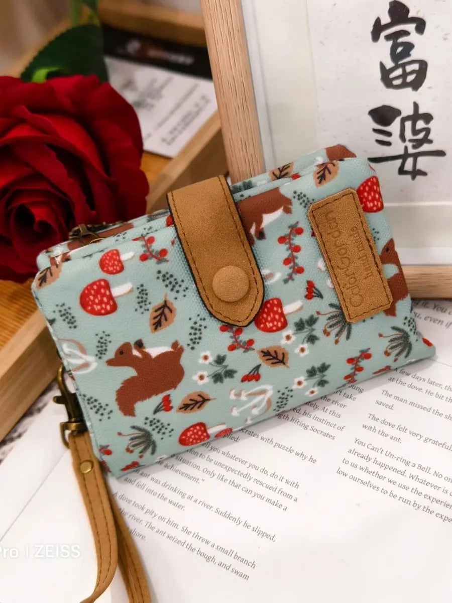 A light blue canvas tri fold multi card wallet featuring a whimsical pattern of brown squirrels red mushrooms and green foliage with tan leather straps and a circular button closure.