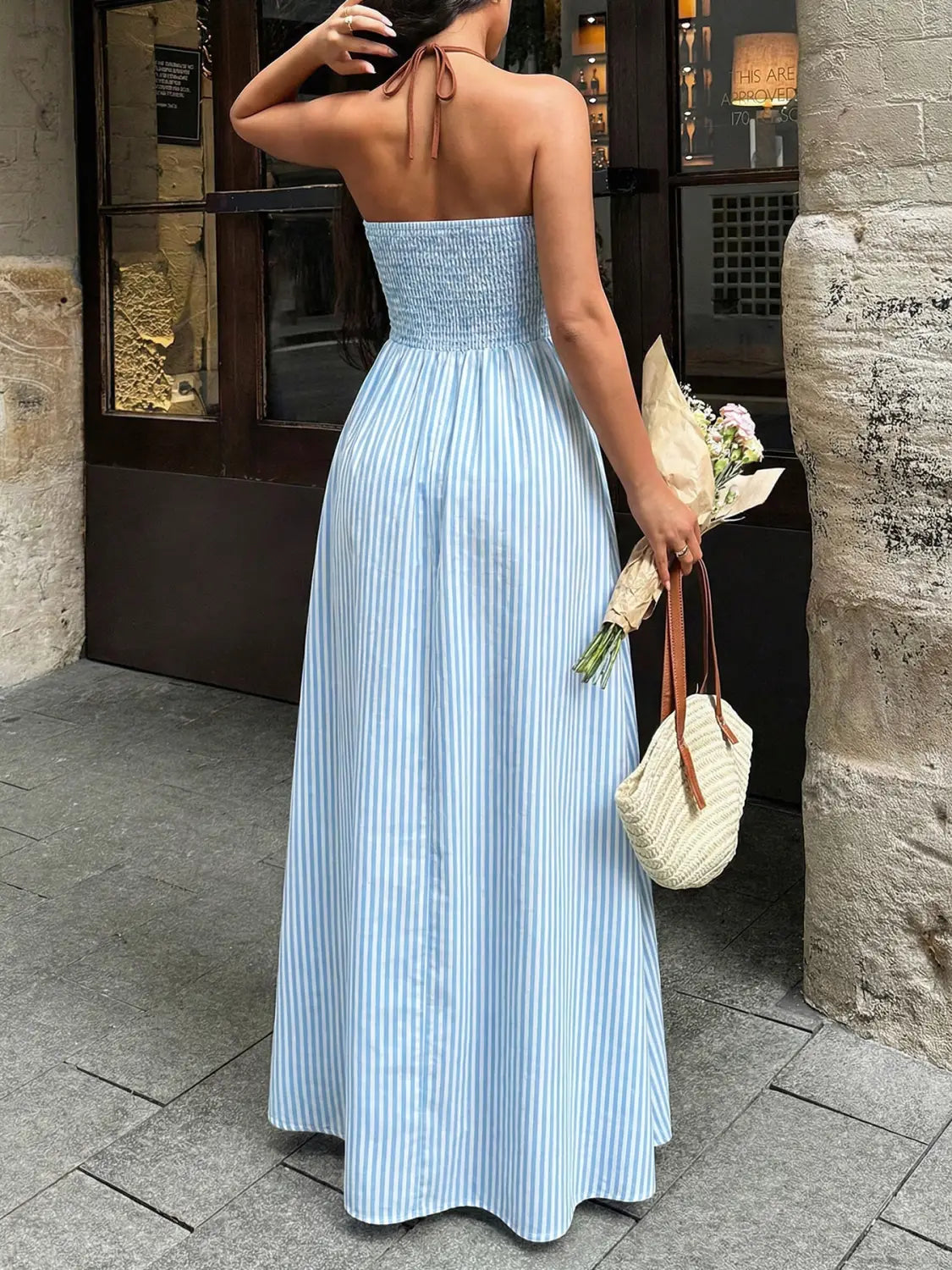 Une robe maxi bustier sans bretelles à rayures verticales bleu clair et blanches, avec un corsage froncé et un design dos noué à encolure en V, idéale pour l’été.