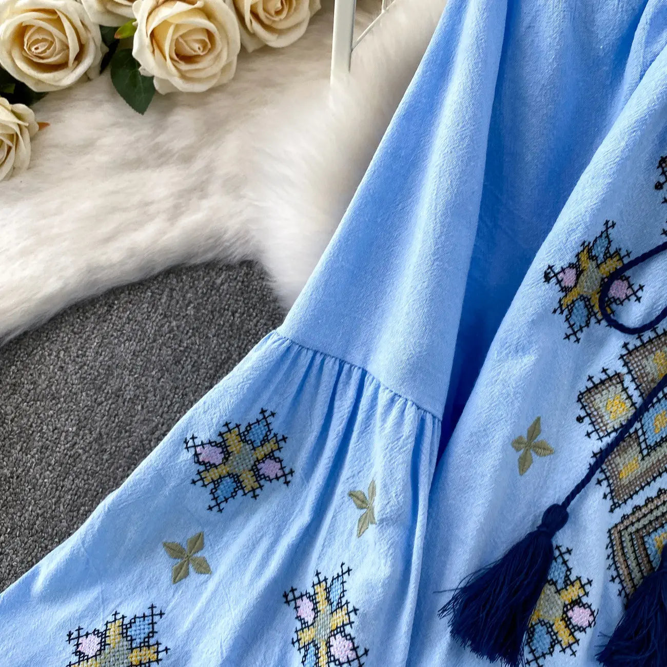 Light blue embroidered lantern sleeve tie neck blouse with intricate yellow pink and beige cross-stitch patterns accented by dark blue tassels.